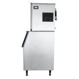 A cube-style ice maker featuring a stainless steel exterior with black trim and four legs. The top section includes a vent and control panel, while the larger bottom section has a slanted, black-edged door for easy ice dispensing.