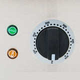 A close-up of a temperature control knob set to 0, with markings from 120 to 570 degrees, on a stainless steel griddle. Next to it, two indicator lights—green and orange—are both on, all set in a metallic panel background.