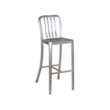 The Anna Barstool features a tall, square seat and curved backrest with three vertical slats. Crafted from brushed aluminum, its minimalist industrial design suits both outdoor/indoor spaces. Footrests connect the straight legs. Plain white background.