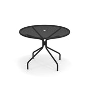 The Cambi Table is a round mesh table with four slender, angled legs curving outward to a central support. Its minimalist, modern outdoor table design features a central hole for an umbrella, and it sits elegantly on a white background.
