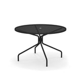 The Cambi Table is a round, black metal outdoor/indoor table featuring a mesh top and four curved, angled legs. With a minimalist design and central umbrella hole, it casts subtle shadows on a plain white background.
