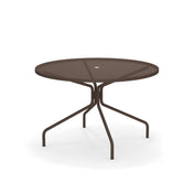 The Cambi Table features a round, brown metal frame with a mesh top and central umbrella hole. Its four slender, curved legs offer stability, making it perfect for outdoor/indoor spaces. The plain white background highlights its sleek design.