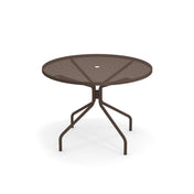 The Cambi Table is a round, metal outdoor table with a mesh top and four angled legs, all in dark brown. It features a small circular hole in the center for an umbrella and is set against a plain white background.