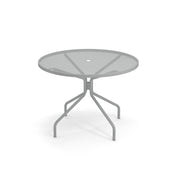 A round outdoor table with a mesh top and a central hole for an umbrella. Its four angled legs curve outward and then downward, ensuring stability on any patio. The background is plain white for a clean, classic look.