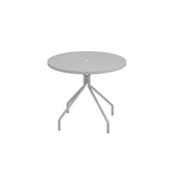 A round, light gray outdoor table with a central hole, supported by four angled legs branching from a single stem. This minimalistic piece is made in Italy, featuring clean lines and a smooth tabletop against a plain white background.