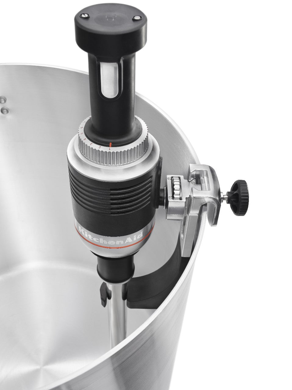 Close-up of a KitchenAid Commercial Immersion Blender with a 12 inch stainless steel blending arm, attached to the rim of a large pot. Secured with a clamp and knob, its control dials and display sit atop the metal heating wand submerged in the pot.