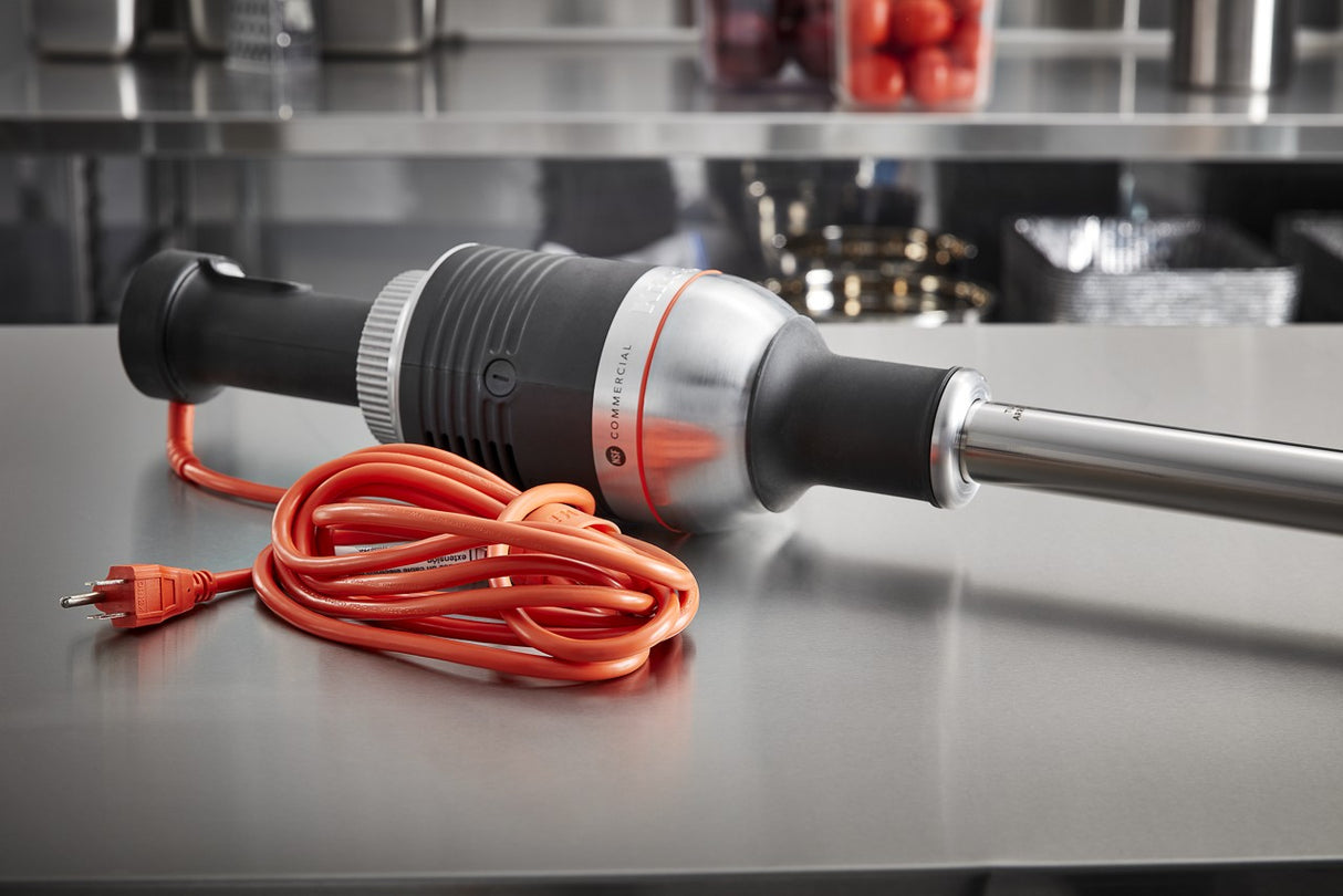 A heavy duty immersion blender with a metallic body, black handle, and long blending shaft lies on a stainless steel kitchen counter. Its bright orange power cord is coiled beside it, with kitchen equipment and shelves visible in the background.