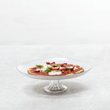A Fortessa Jupiter clear cake stand holds a small pizza topped with melted cheese, tomato slices, pepperoni, and fresh basil, set against a light gray textured background. The scene is minimalistic with no other objects present.