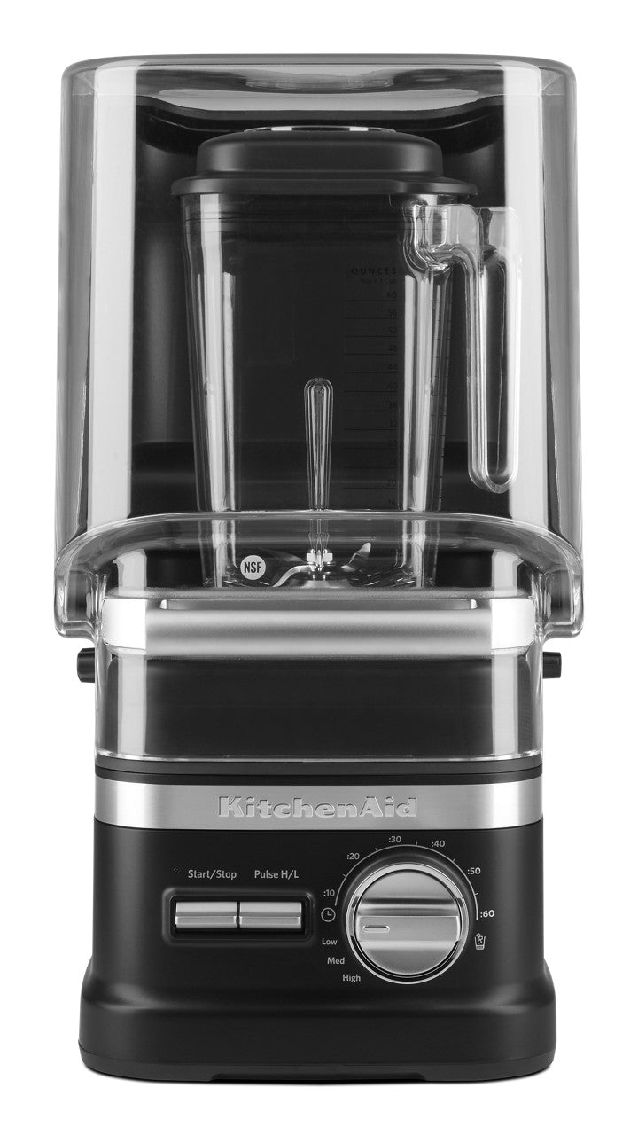 A black and silver KitchenAid Commercial Blender is shown with a clear plastic sound enclosure, a transparent 60 oz blending jar with handle, and control knobs and buttons. The KitchenAid logo is visible on the base below the controls.