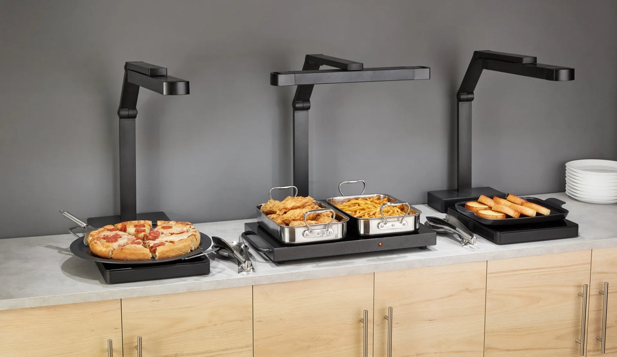 Three buffet-style food warmers with NSF-certified multi-position heat lamps sit on a countertop: sliced pizza, fried chicken and French fries, and bread rolls. Stacks of white plates are on the far right against a gray wall.