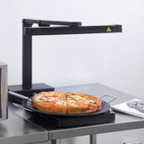 A whole deep-dish pizza rests on a black warming tray beneath an NSF-certified infrared heat lamp on a stainless steel countertop in a commercial kitchen. A pizza server and a folded white cloth are conveniently placed nearby.