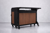 The NomadBar Mobile features a black metal frame, vertical wooden slats, four caster wheels, a black countertop, and an insulated ice bin. Four upside-down stemmed glasses hang just below the top. The background is plain and light gray.