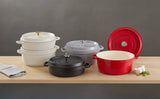 Five Ironlite Casserole enameled cast iron Dutch ovens in white, gray, black, and red sit on a wooden table. Perfect for induction cooking, these non-stick casseroles are paired with garlic, herbs, and a towel for added charm.