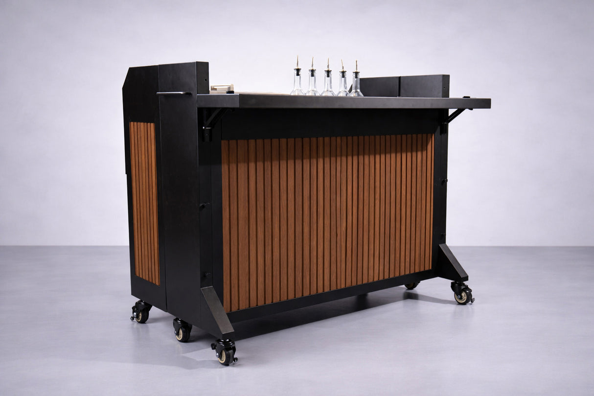 A NomadBar Mobile portable bar cart with a dark frame, vertical wooden slats on the front, and four caster wheels. On the bar top are five clear glass bottles with pour spouts, a small wooden block, and an insulated ice bin.