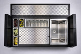 A top-down view of a sleek, modern bar station with a stainless steel sink, six glass bottles, metal containers of garnishes, and an insulated ice bin. The NomadBar Mobile features perforated metal panels in a black frame against a light background.