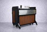 A modern NomadBar Mobile collapsible bar system with a black metal frame, wood paneling, perforated metal front, and caster wheels. On top are liquor pourers, a jigger, and bar tools—all set against a plain gray backdrop.