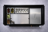 Overhead view of a sleek, modern NomadBar Mobile station with metal compartments holding cherries, olives, lime slices, and orange peels, five bottles in holders, an insulated ice bin, grated workspace, and a closed stainless steel cabinet on a gray surface.