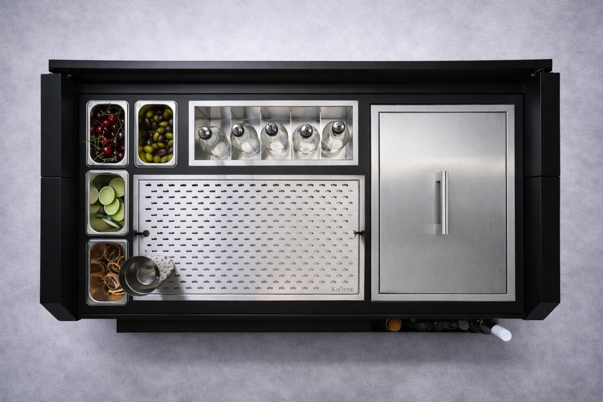 A top-down view of a sleek NomadBar Mobile station with compartments for cherries, olives, lime wedges, and orange slices. Six glass bottles with pour spouts sit beside a perforated workspace and an insulated ice bin in this collapsible bar system.