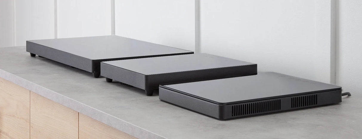 Three rectangular, low profile induction cooktop devices in varying sizes sit side by side on a gray countertop against a white paneled wall. The minimalist, dark gray appliances have ventilation slots on their sides.
