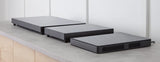 Three rectangular, low profile induction cooktop devices in varying sizes sit side by side on a gray countertop against a white paneled wall. The minimalist, dark gray appliances have ventilation slots on their sides.