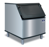 A commercial Manitowoc stainless steel ice bin with a black, slanted hinged lid on top, mounted on four metal legs. The shiny bin features a small manufacturer logo on the front. Perfect for use with a top-mounted ice maker. Background is plain light gray.