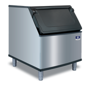 A commercial Manitowoc stainless steel ice bin with a black, slanted hinged lid on top, mounted on four metal legs. The shiny bin features a small manufacturer logo on the front. Perfect for use with a top-mounted ice maker. Background is plain light gray.