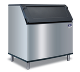 A commercial stainless steel Manitowoc ice bin with a black hinged lid stands on four metal legs against a plain white background. The front features a small blue and white manufacturer’s badge in the upper right corner, ready for a top-mounted ice maker.