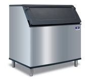 A commercial stainless steel Manitowoc ice bin with a black hinged lid stands on four metal legs against a plain white background. The front features a small blue and white manufacturer’s badge in the upper right corner, ready for a top-mounted ice maker.