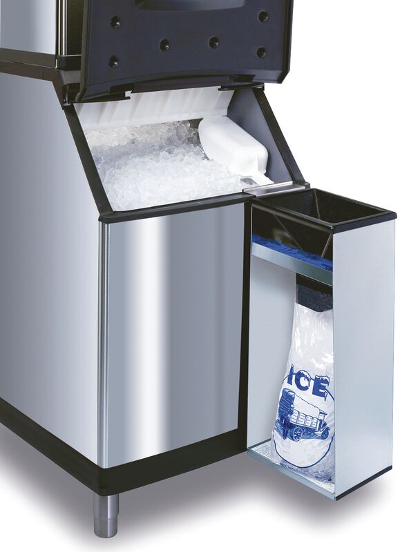 A Manitowoc commercial ice machine with a stainless steel body dispenses ice. The upper compartment is open, revealing ice and a white scoop. A lower side Ice Bagger Assembly holds a clear plastic bag labeled “ICE,” partially filled with cubes.