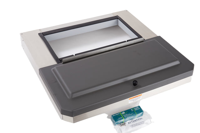 A Manitowoc stainless steel commercial kitchen equipment features an open rectangular top compartment with a hinged black plastic lid. A green label packet marked “Attention” sits in front of the unit on a white surface.