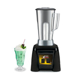 A high-power blender with a stainless steel container and black and yellow base stands on a white background. Next to it is a tall glass filled with green milkshake, topped with whipped cream, a red-striped straw, cherry, and mint sprig.