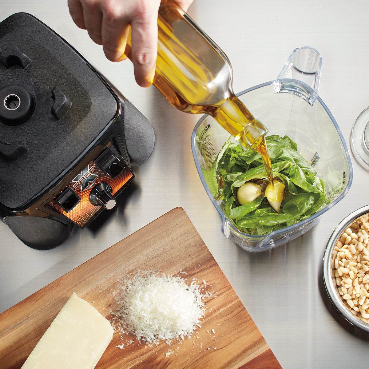 A hand pours olive oil into a BPA Free Blender pitcher with spinach and garlic. Nearby, Parmesan—both grated and in a block—and a bowl of pine nuts rest on a board, while the blender base sits on the metal countertop.