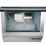 A commercial ice maker filled with clear, rectangular ice cubes sits beneath a white plastic scoop. The stainless steel front features the MAXX ICE logo in the lower right corner, and an ample ice storage bin ensures plenty of supply.