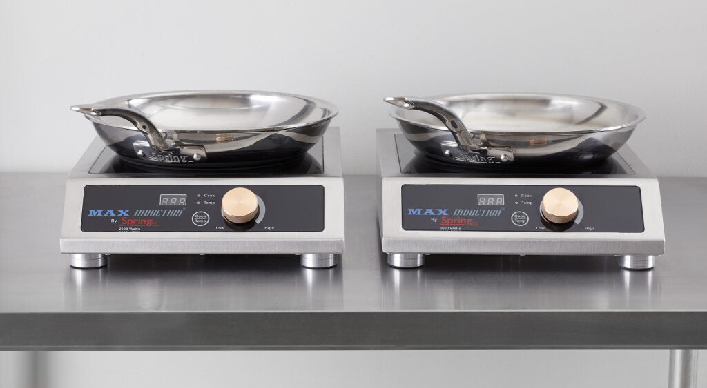 Two stainless steel induction cooktops with digital displays and large dials sit side by side on a metal countertop. Each features a stainless steel fry pan with an aluminum core, handles pointing outward, against a plain wall.