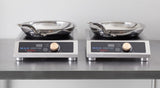 Two stainless steel induction cooktops with digital displays and large dials sit side by side on a metal countertop. Each features a stainless steel fry pan with an aluminum core, handles pointing outward, against a plain wall.