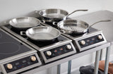 Four stainless steel frying pans with aluminum core sit on four separate induction cooktops atop a metal table. Each cooktop features digital displays and control knobs. One pan has a ridged grill surface, all set against a plain white background.