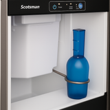 A blue bottle stands upright inside a Scotsman Nugget ice maker, resting against a metal holder. The clean, empty interior highlights the control buttons and indicator lights on the top front panel of the nugget ice machine.