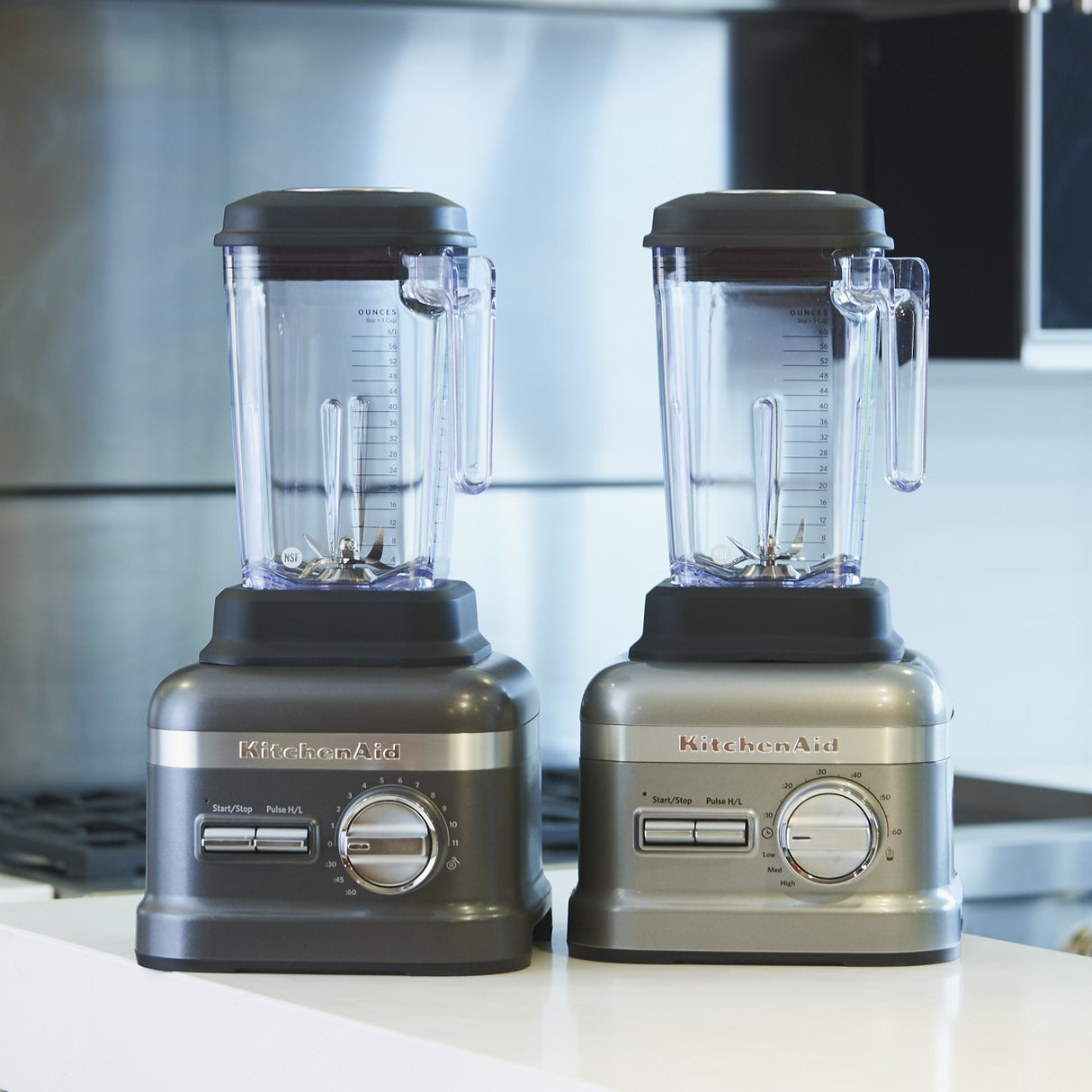 Two silver KitchenAid Commercial Bar Blenders with clear 60 oz pitchers and Patented Talon blades sit side by side on a white kitchen counter, blending seamlessly into a modern kitchen with stainless steel appliances.