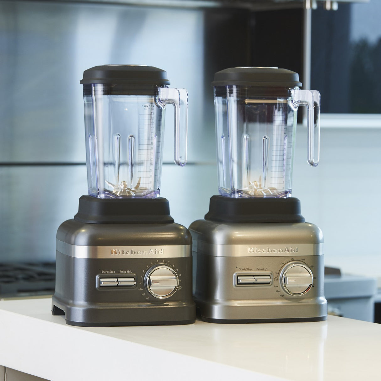 Two metallic KitchenAid Commercial Bar Blenders with clear 60 oz. capacity pitchers and black lids sit side by side on a white kitchen counter. Both feature control knobs and the patented Talon asymmetrical blade for efficient blending.