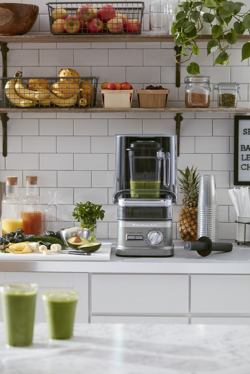 Modern kitchen with a silver KitchenAid Commercial Blender filled with green smoothie on a white counter. Two green smoothies in glasses are in front, surrounded by fresh fruits, leafy greens, juice jars, and kitchen containers against a white tile backsplash.