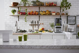 A bright kitchen with a marble counter holds two green smoothies made with a BPA free blender. Behind it are white cabinets, a juicer, knives, and assorted fruits and vegetables on wooden shelves. Potted plants and a menu board decorate the white-tiled backsplash.