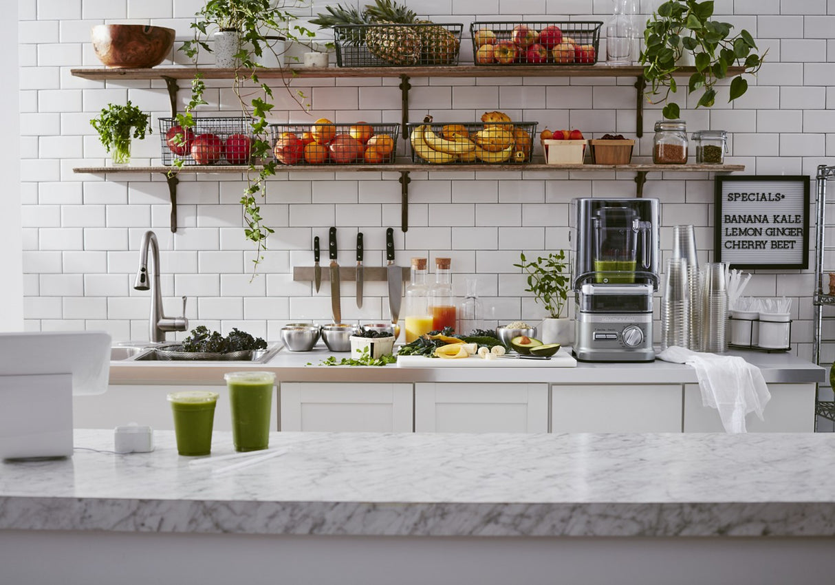 A clean, modern kitchen with a marble counter features two green smoothies. Nearby, a BPA free blender sits ready for use. Open shelves display fresh fruits, leafy greens, and potted plants, with a sign listing smoothie ingredients by the white-tiled wall.