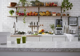 A clean, modern kitchen with a marble counter features two green smoothies. Nearby, a BPA free blender sits ready for use. Open shelves display fresh fruits, leafy greens, and potted plants, with a sign listing smoothie ingredients by the white-tiled wall.