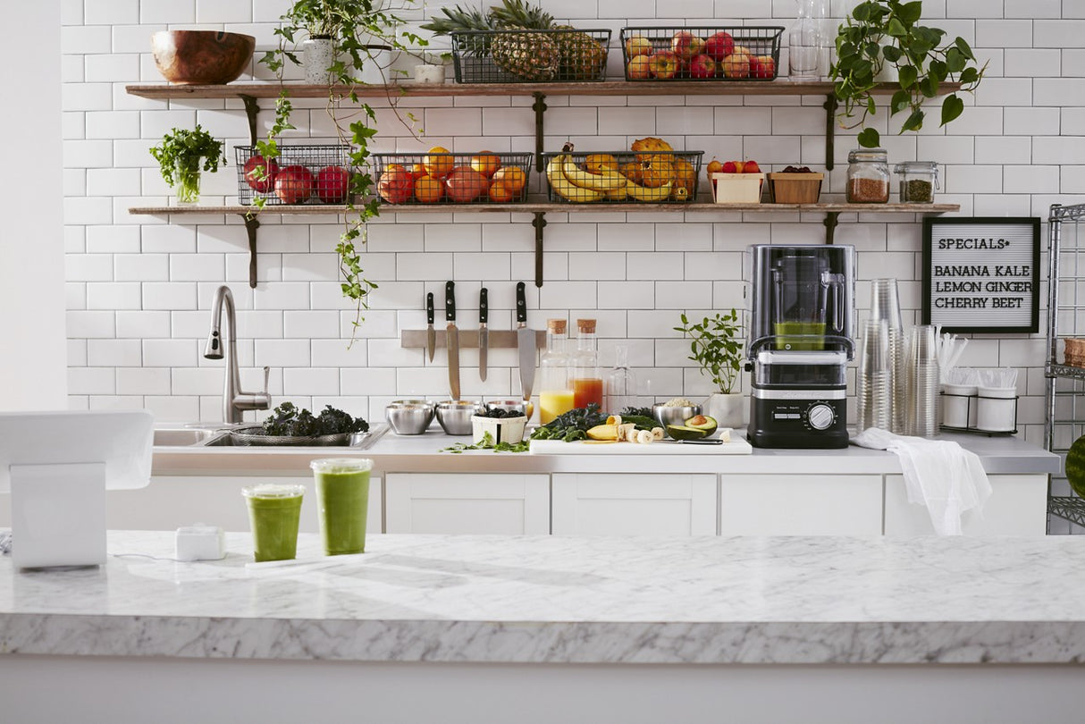 A bright kitchen with white subway tiles, open shelves holding assorted fruits, bowls, and potted plants. A KitchenAid Commercial Blender with 60 oz. capacity sits on the counter near fresh ingredients and two green smoothies on a marble island.