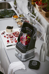 A BPA free blender with a clear sound shield is filled with chopped red fruit. It sits on a kitchen counter beside a cutting board with cut pomegranate, scattered berries, stacked clear cups, a dish towel, and baskets of fresh fruit.