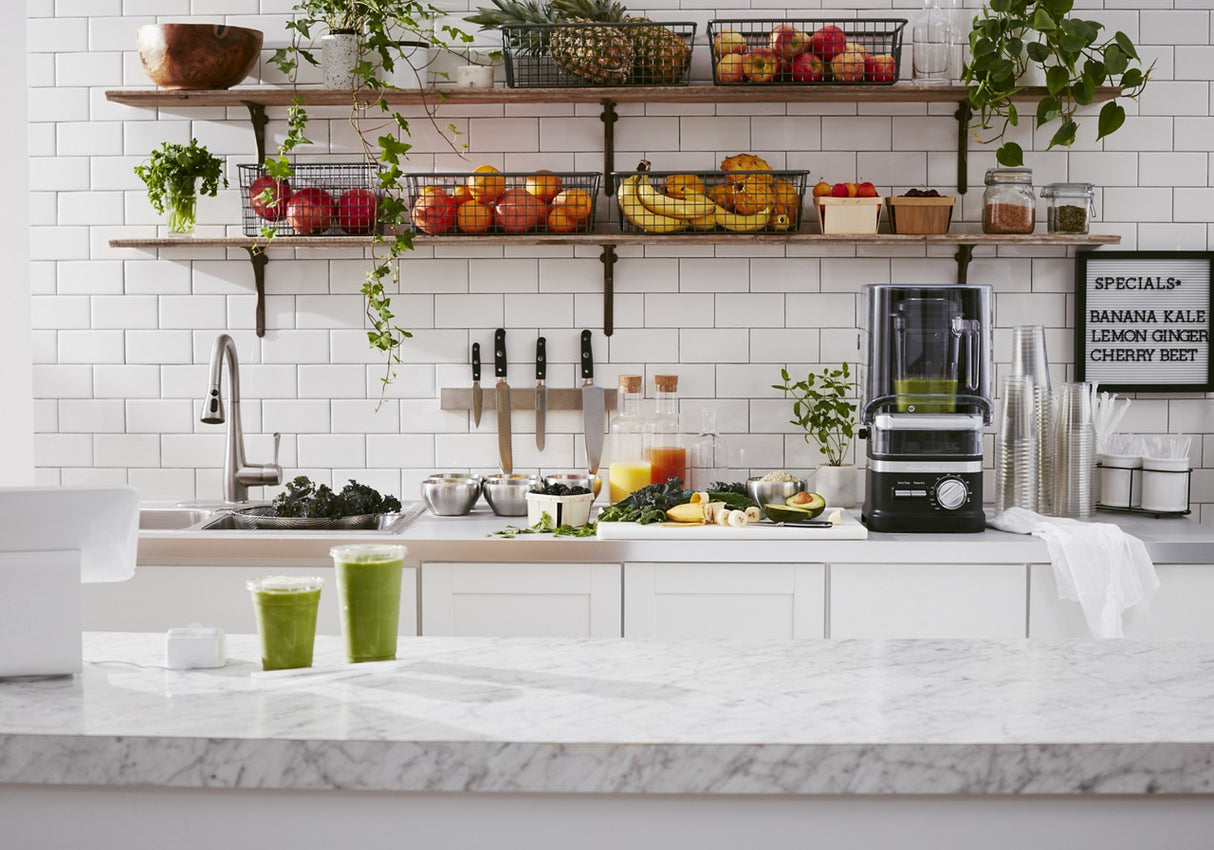 A bright, modern kitchen features a marble counter with two green smoothies. Next to the sink, a 60 oz blender stands ready. Shelves display fruit, plants, and jars, while a letter board lists smoothie flavors against classic white subway tiles.
