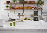 A bright, modern kitchen features a marble counter with two green smoothies. Next to the sink, a 60 oz blender stands ready. Shelves display fruit, plants, and jars, while a letter board lists smoothie flavors against classic white subway tiles.