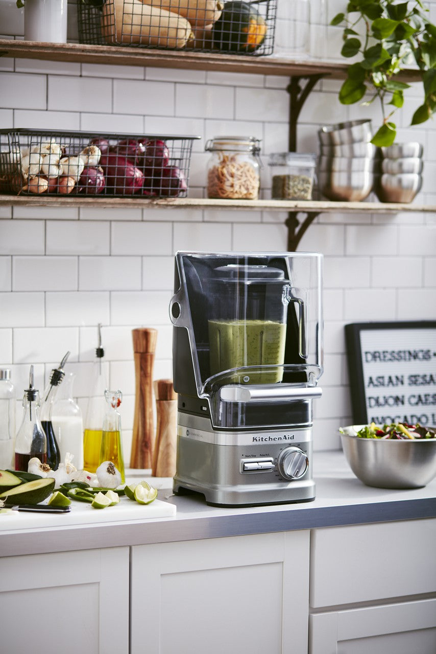A silver KitchenAid Commercial Blender with a clear pitcher, filled with green smoothie, sits on a white kitchen counter. Nearby are bottles of oil, chopped vegetables, and a salad bowl. Overhead shelves hold baskets with onions and jars, plus hanging plants.