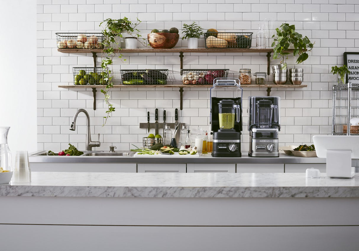 Modern kitchen with a white tile backsplash, marble countertop, and open shelves holding produce and plants. A KitchenAid Commercial Blender with 60 oz. capacity sits by the sink among knives and vegetables in this clean, bright, organized space.