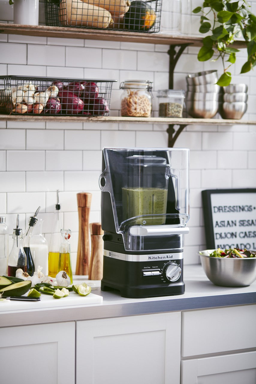 A KitchenAid Commercial Blender with a 60 oz. capacity and clear sound shield is blending a green smoothie on a modern kitchen counter. Sliced cucumbers, limes, and baskets of onions and potatoes add to the fresh kitchen scene in the background.